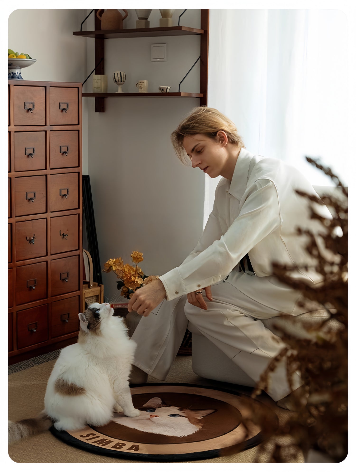 Woman in a white outfit interacting with a cat on a rug in a room with wooden drawers and a shelf.